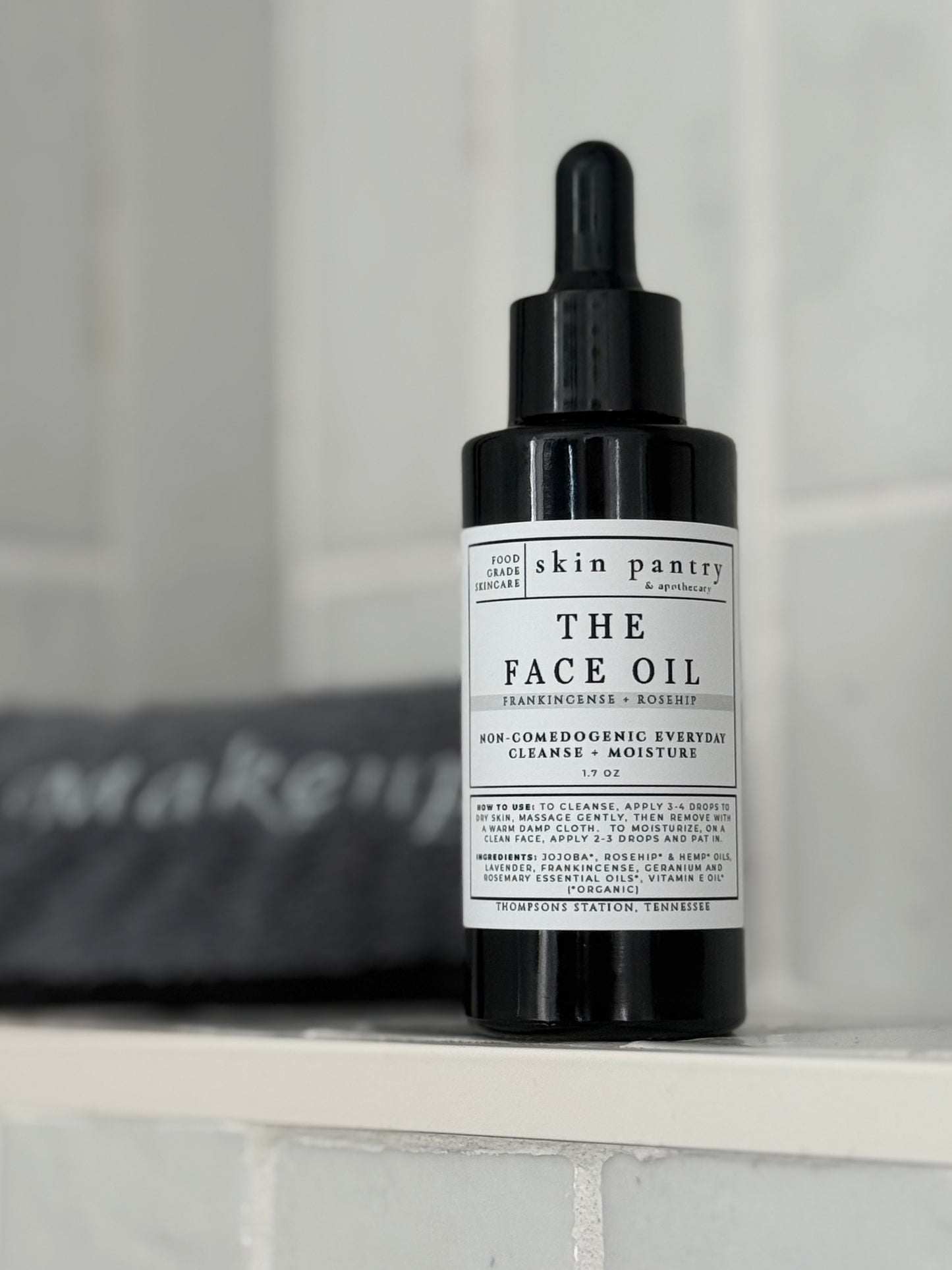 A black dropper bottle of Skin Pantry 'The Face Oil' with a white label, resting on a white shelf against a soft gray background.